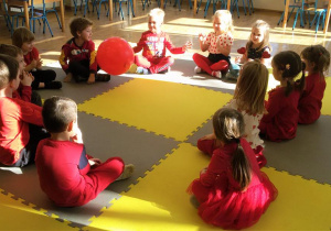 Widok na siedzące w kręgu dzieci, które podają sobie duży, czerwony balon.